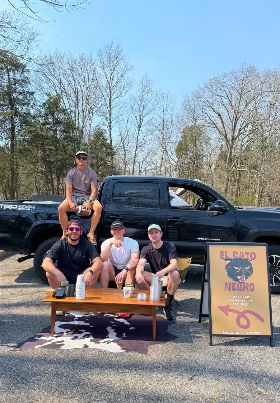 Four people around a mid-century coffee table and cowhide rug set up on the pavement beside a truck at R.A.D. 100, with the El Gato Negro sandwich-board sign